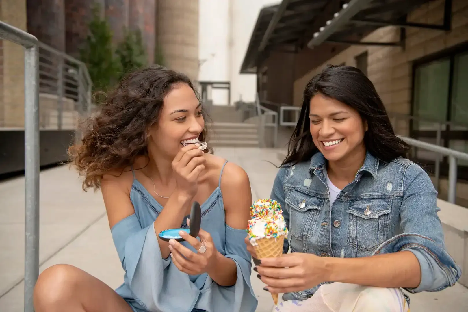 Two women are sitting on the sidewalk eating ice cream cones.