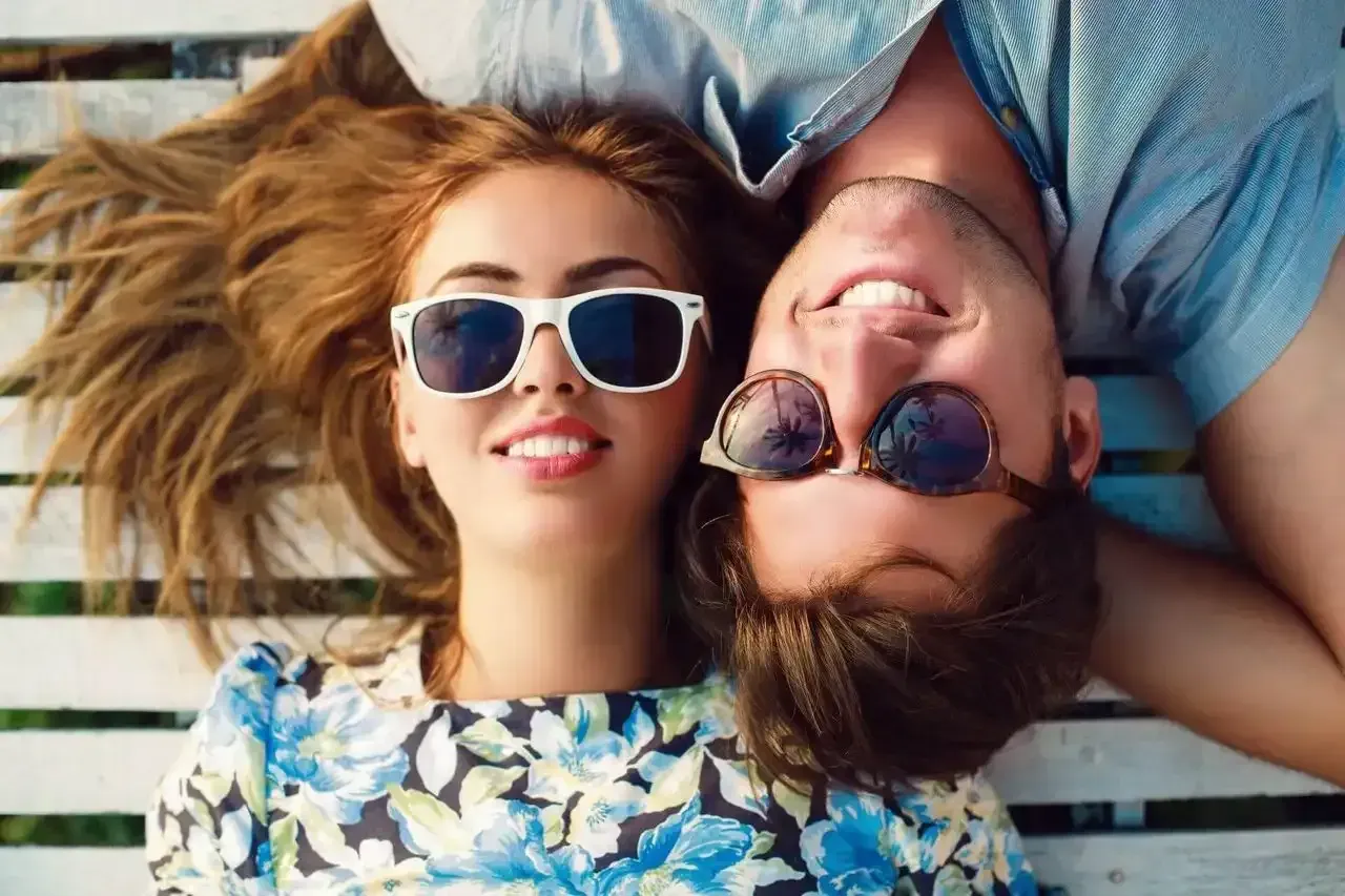 A man and a woman wearing sunglasses are laying on a bench.