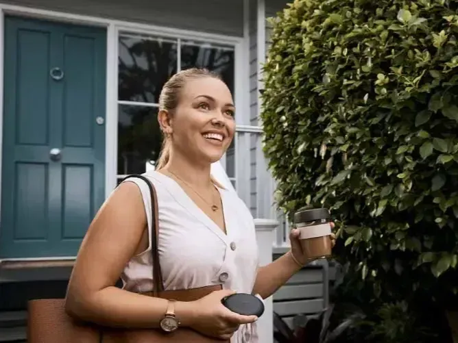Woman smiling outside a teal door, holding coffee and a bag. She has keys in her hand and is near green foliage.