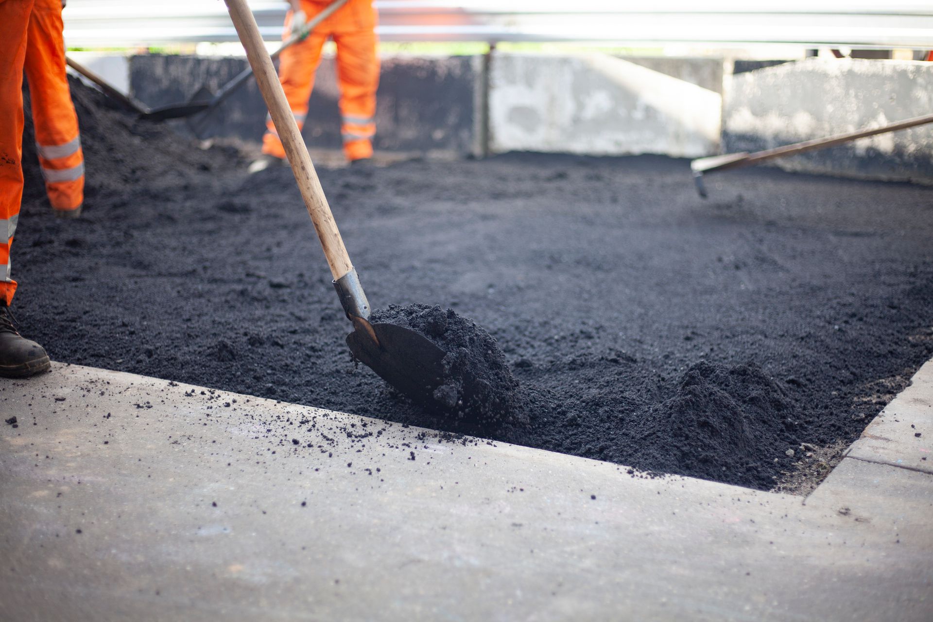 Workers in orange overalls spreading asphalt with shovels and rakes on a road surface.