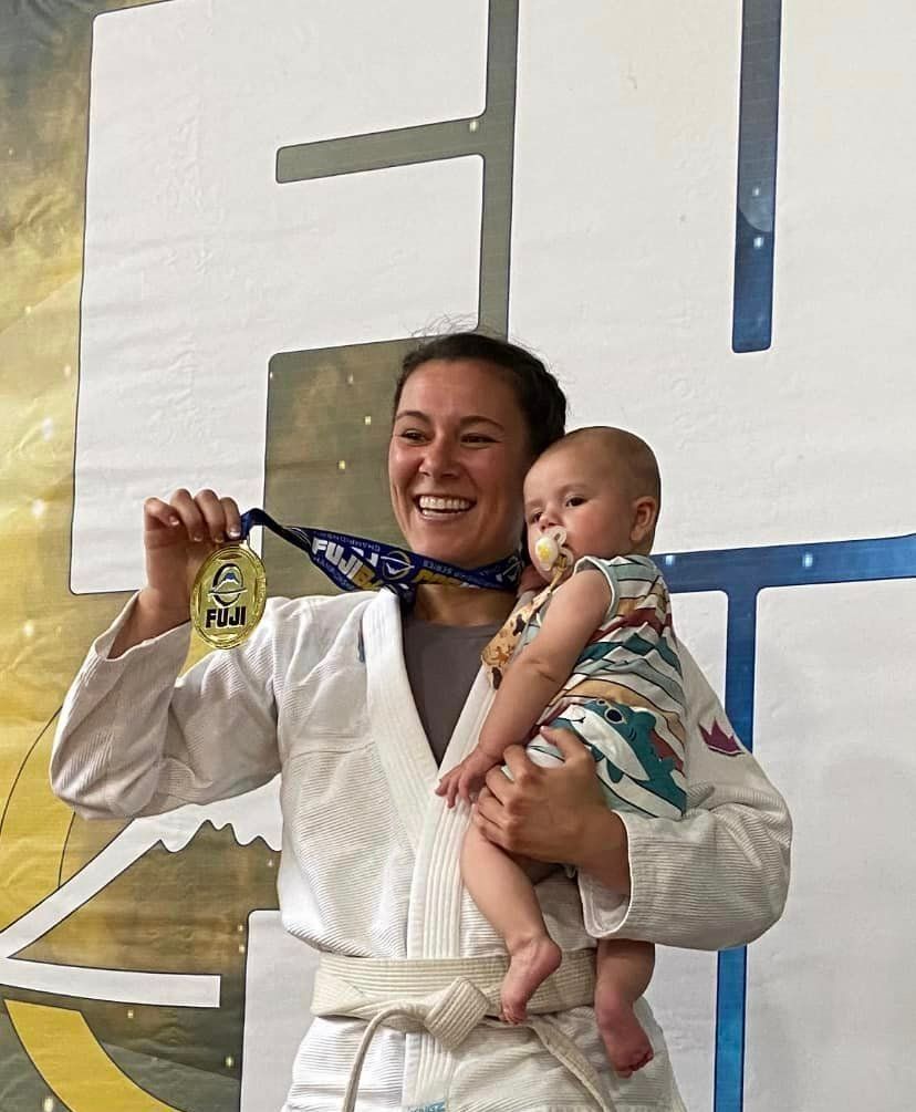 Woman in white gi holds baby and gold medal, smiling on a podium.