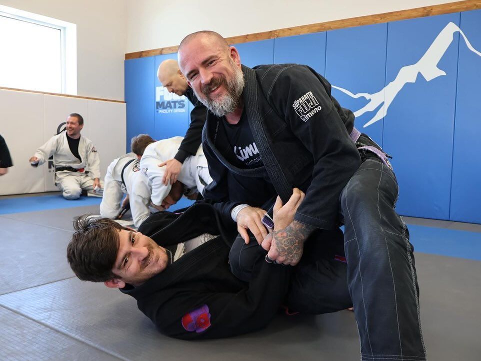 Man smiling over another in a jiu-jitsu match; both in black gis, on blue mat in a gym. Other practitioners in background.