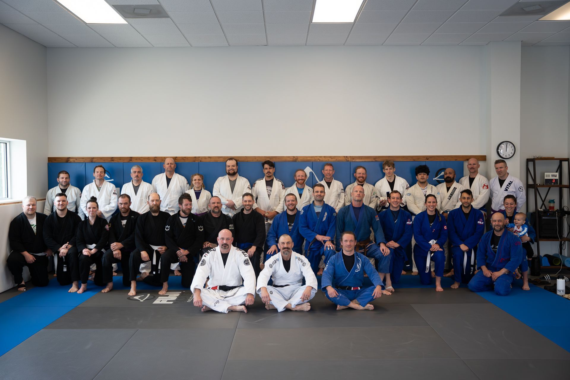 Group photo of a Jiu-Jitsu class in a dojo. People wearing various gi colors pose together.