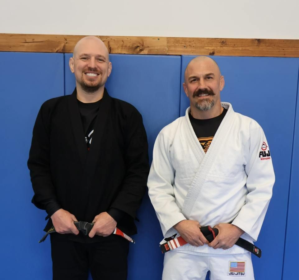 Two bald men in martial arts attire stand in front of a blue wall. One wears black, the other a white gi.