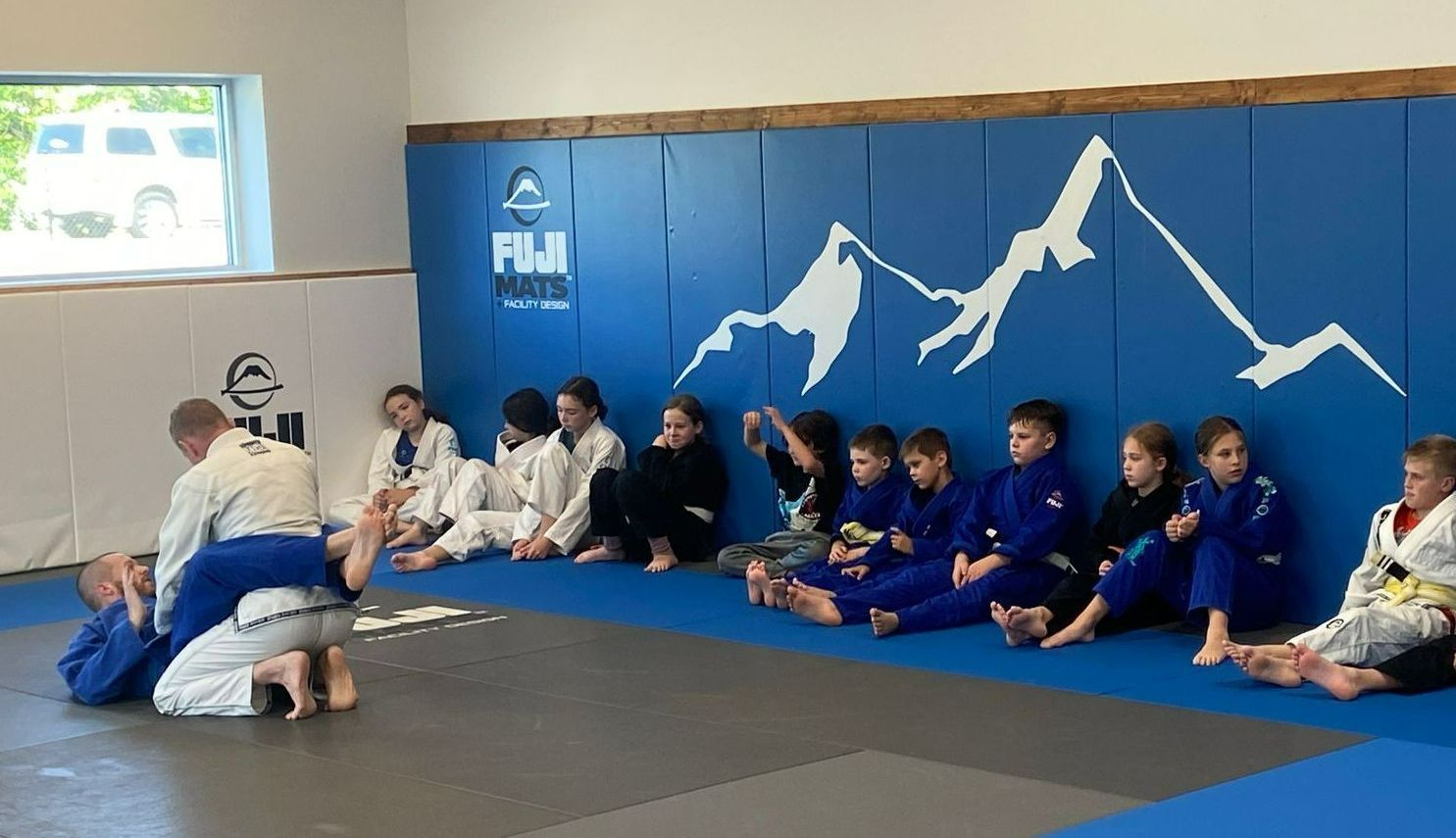 Kids in blue and white gis watch a Jiu-Jitsu demonstration on a mat in a gym.