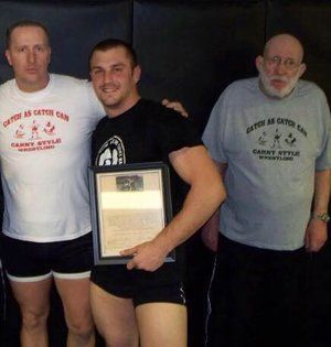 Three men pose in a gym. Center man holds a framed paper, wearing black shorts and a black shirt.