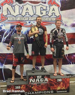 Three men on a podium at a NAGA tournament. The first place winner holds a belt.