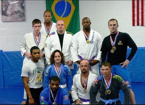 Group of people in martial arts attire with medals, standing in front of a Brazil flag.