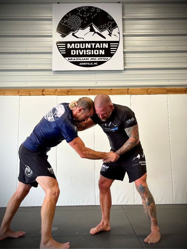 Two men in a grappling hold on a mat at a gym. Sign reads 