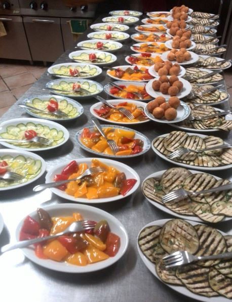 Un bancone da cucina professionale, fiancheggiato da numerosi piatti con pietanze già pronte, tra cui verdure affettate e polpette fritte.