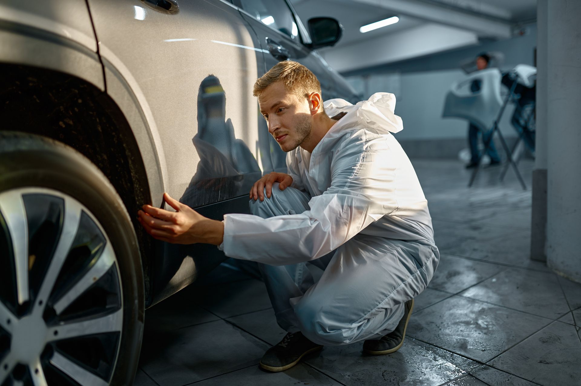 A man in a white suit is kneeling down next to a car in a garage.