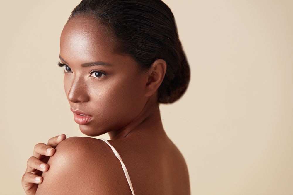 Woman with dark skin looks over her shoulder, touching her shoulder. Beige background.