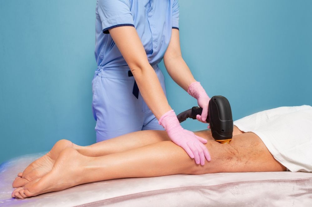 Person in blue scrubs uses a laser device on a person's leg lying on a table. Pink gloves and a blue backdrop.