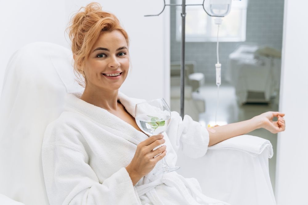 Woman in white robe, with IV drip, smiling and holding a drink in a bright room.