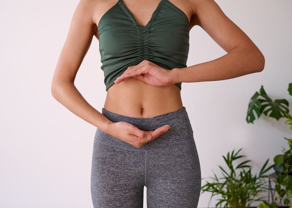 Woman in athletic wear holding hands near abdomen, against white wall and plants.