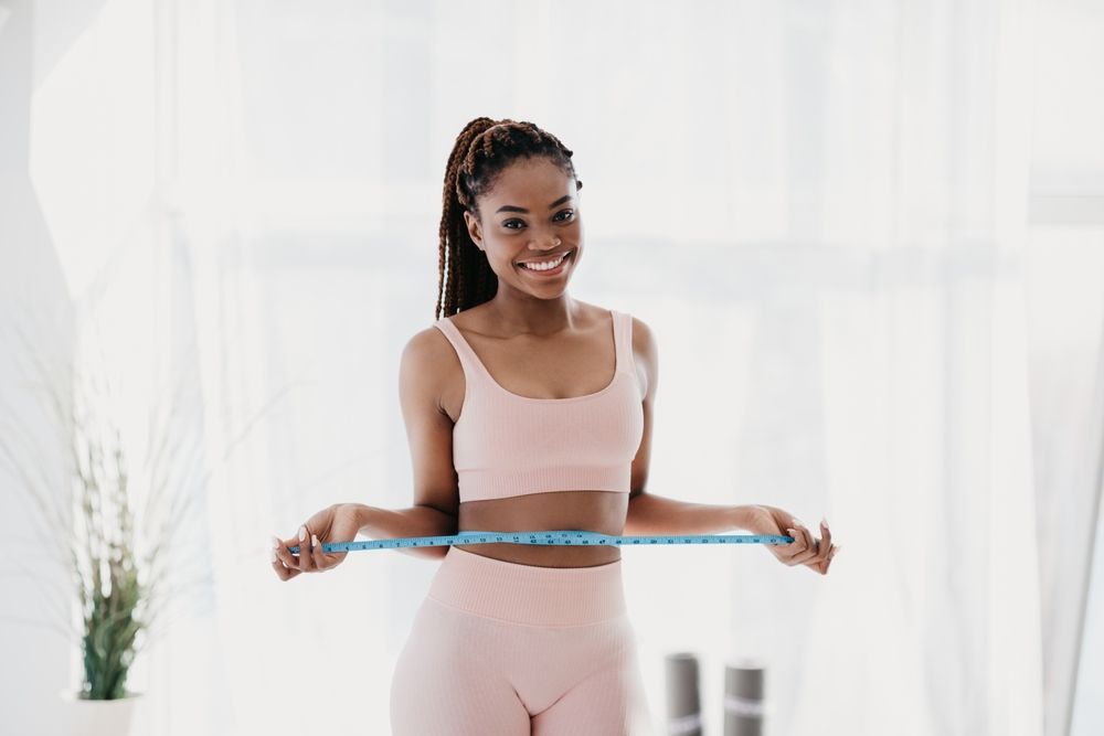 Woman in pink activewear measures waist with a blue tape, smiling in a bright room.