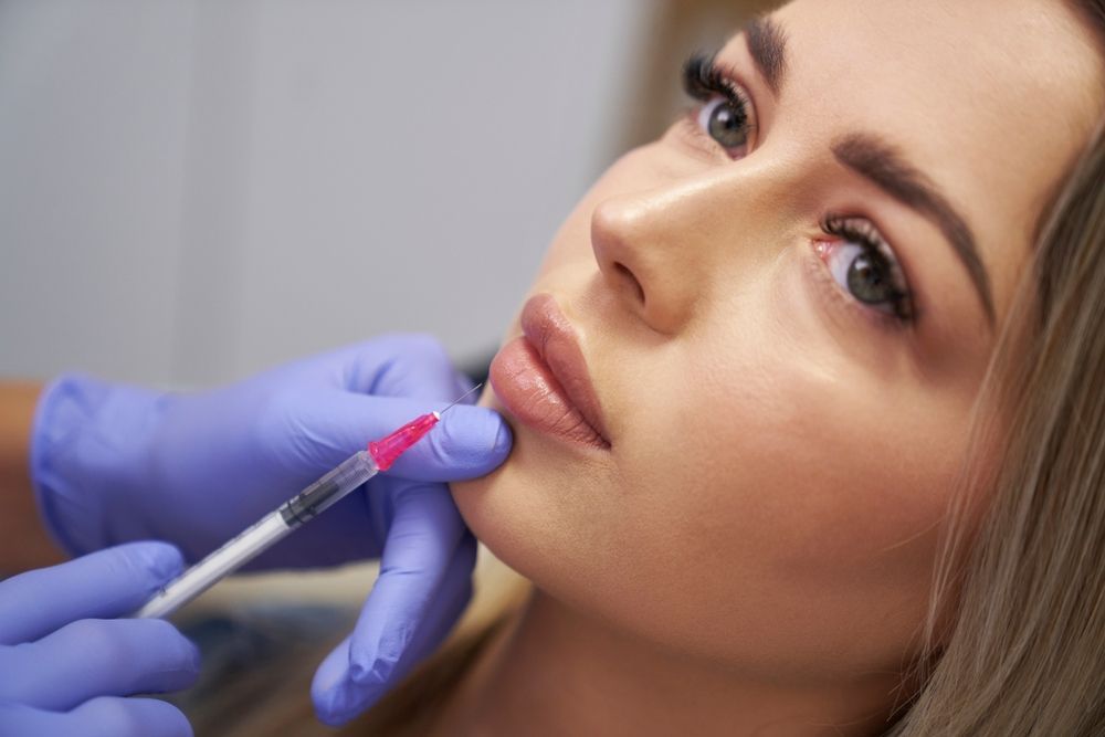 Person receiving lip filler injection. A gloved hand holds a syringe near the lip.