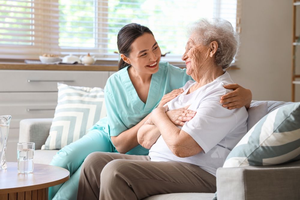 Caregiver smiles, comforting elderly person sitting on couch; living room.