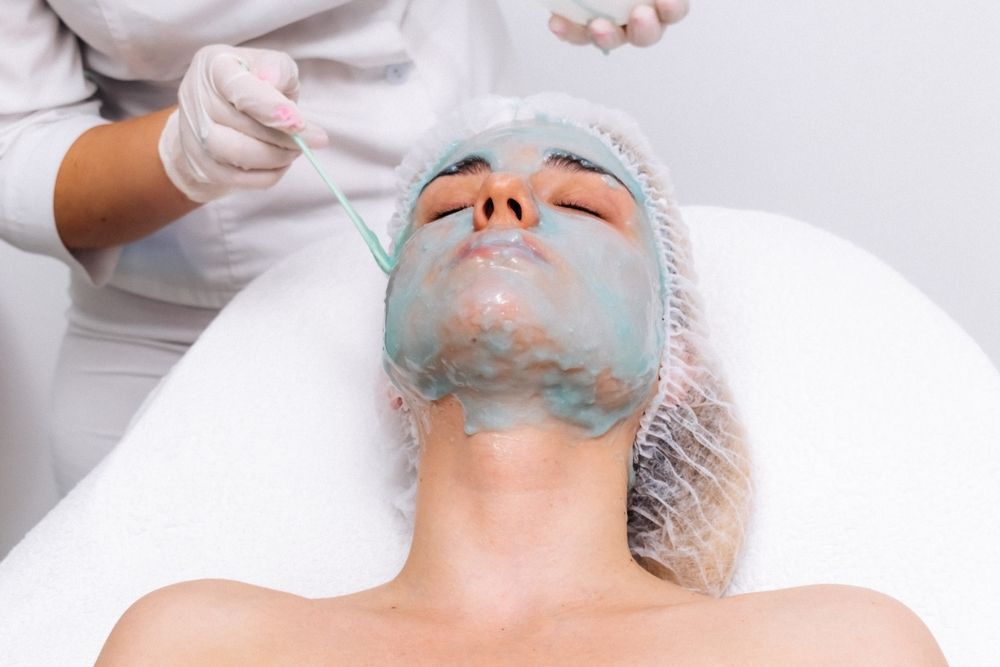 Woman getting facial treatment; esthetician applying blue mask in a spa setting.