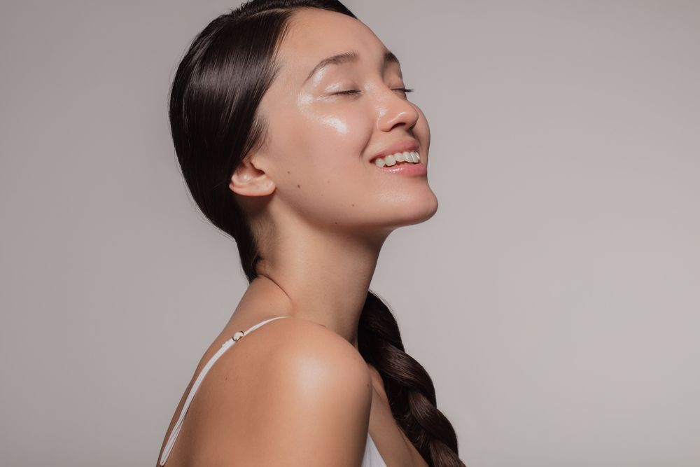 Woman with dark hair in a braid smiles, eyes closed, with glowing skin, against a neutral backdrop.