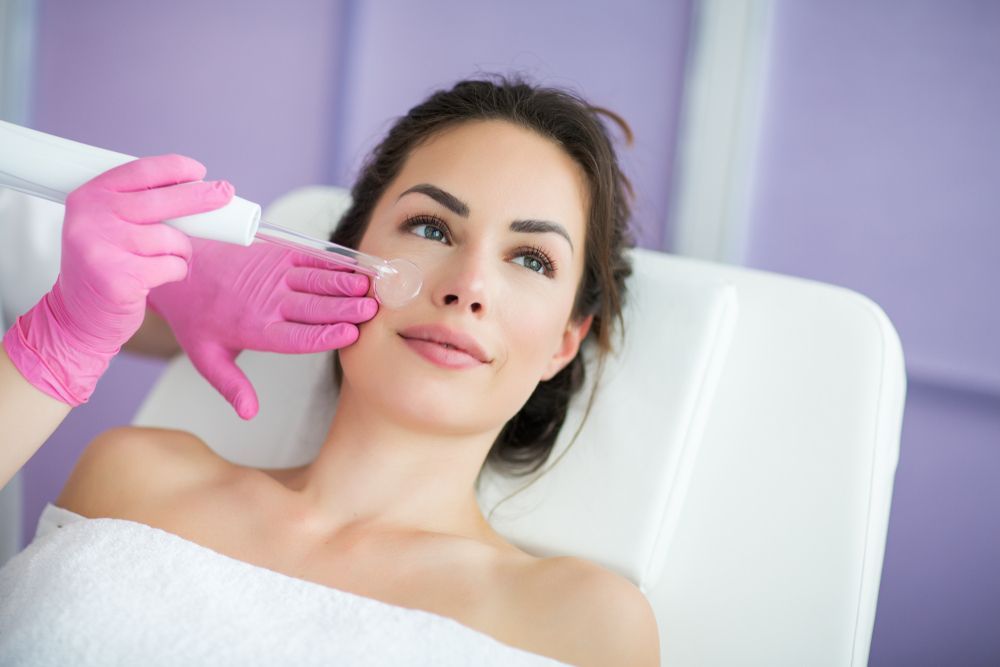 Woman receiving facial treatment in a clinic; pink gloves, tool on face, white towel, purple wall.