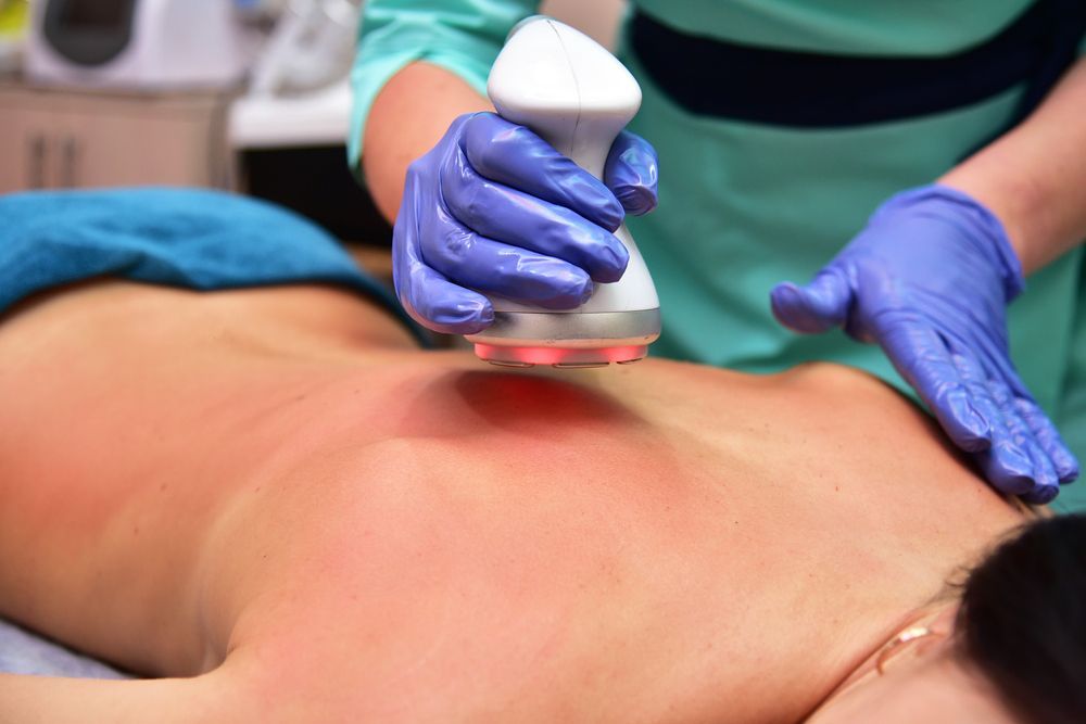 Person receiving laser therapy on their back, red light visible. Therapist's gloved hands hold device.
