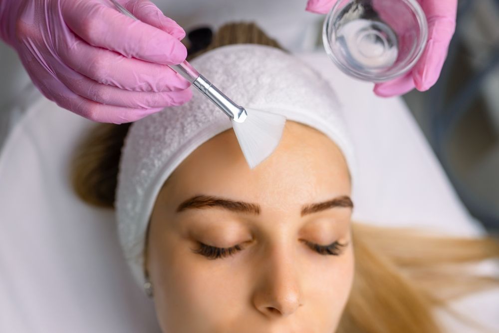 Person receiving chemical peel treatment on forehead with brush. Pink gloves, white headband, clear liquid.