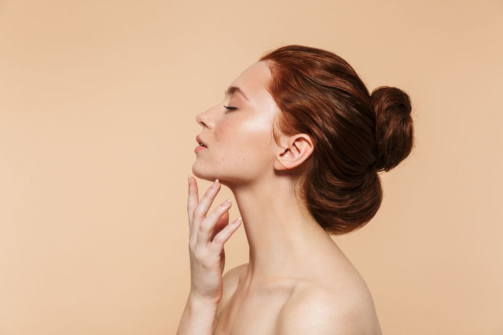 Woman with red hair in a bun, hand on her neck, eyes closed, against a beige background.