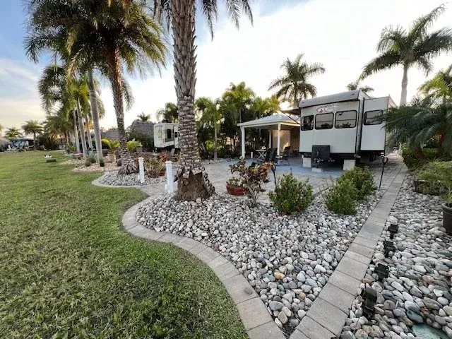RV parked in a tropical landscape, featuring palm trees, gravel, and a paved seating area.