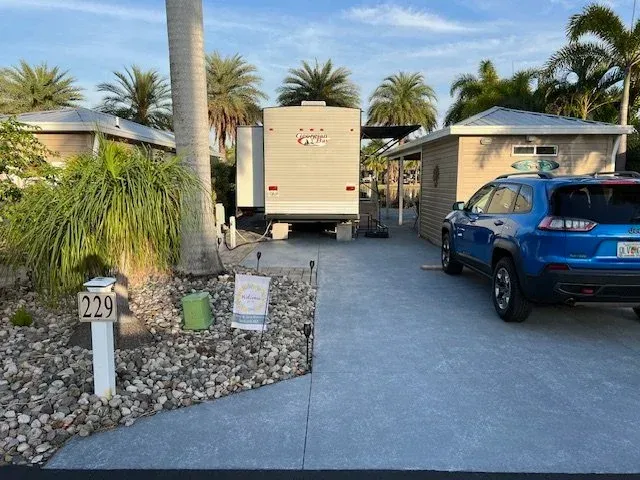 RV parked in a campsite with a blue SUV, small buildings, and palm trees under a blue sky.