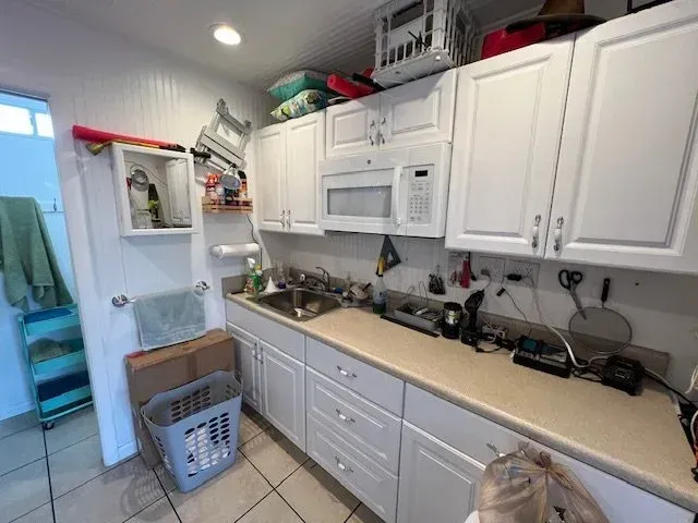 Small white kitchen with upper and lower cabinets, microwave, and laundry hamper.