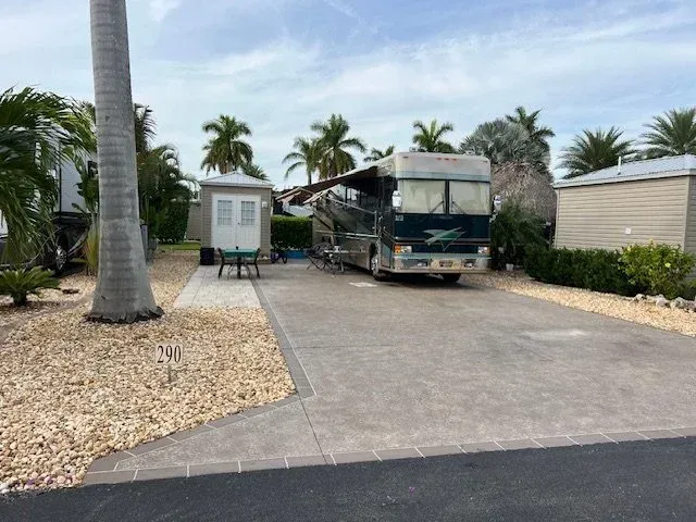A large RV parked on a paved lot, palm trees, small white building, and stone landscaping.