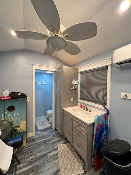 Bathroom with blue walls, gray vanity, and a ceiling fan. Toilet visible through doorway.