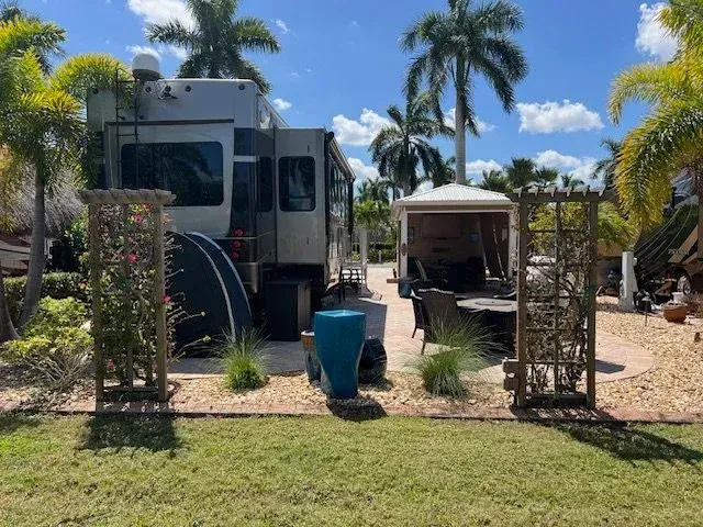 RV parked on a patio with a gazebo, palm trees, and a blue water fountain in a sunny outdoor setting.
