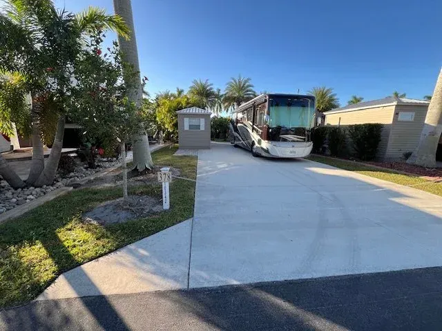 RV parked in concrete RV park space. Palm trees and small utility building visible. Sunny day.