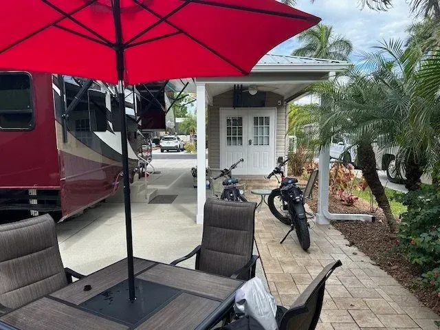 Red umbrella shades patio furniture next to a motorhome and a small building with white doors.