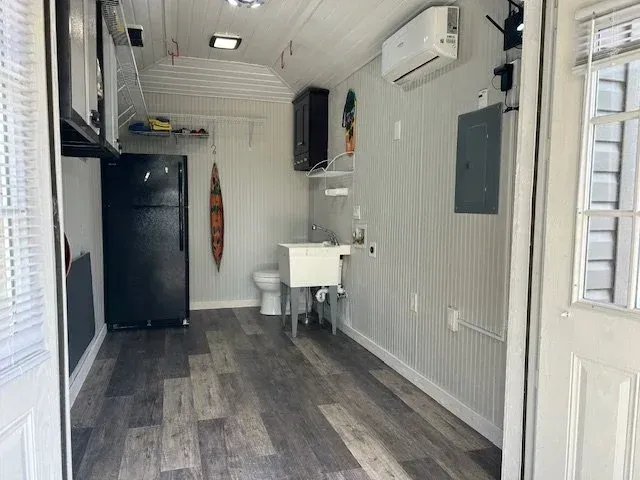 Interior of a tiny home with a kitchen, bathroom, and fridge; light gray walls, wood floor.