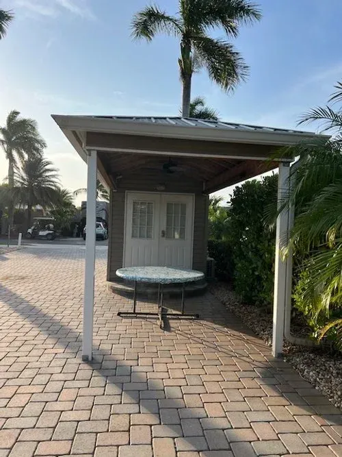 Pavilion with a round table, doors, and palm trees; brick pavement.