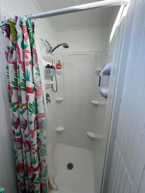 Small white shower with a tropical print curtain, showerhead, and built-in soap shelves.