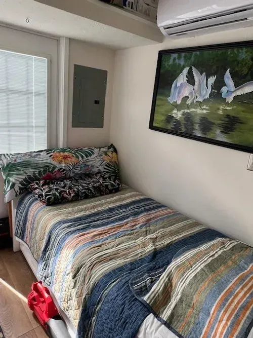 A small bedroom with a twin bed, a painting, and a window. The bed is made with colorful bedding.