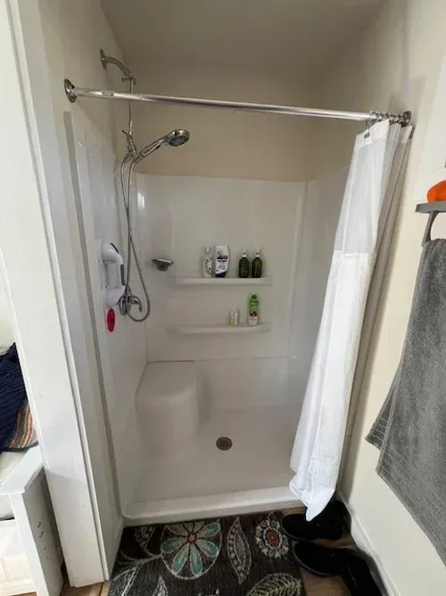 White accessible shower stall with built-in seat, shelves, and chrome shower head. Open shower curtain.