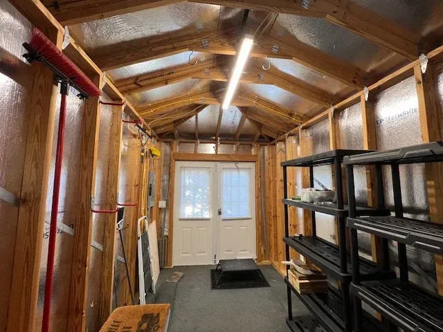 Interior of a shed with wooden beams, shelving, and a door at the end. Red broom leaning against the wall.