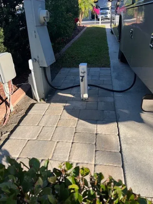 RV site with gray paving, electrical hookup, and RV parked next to grass and shrubs.