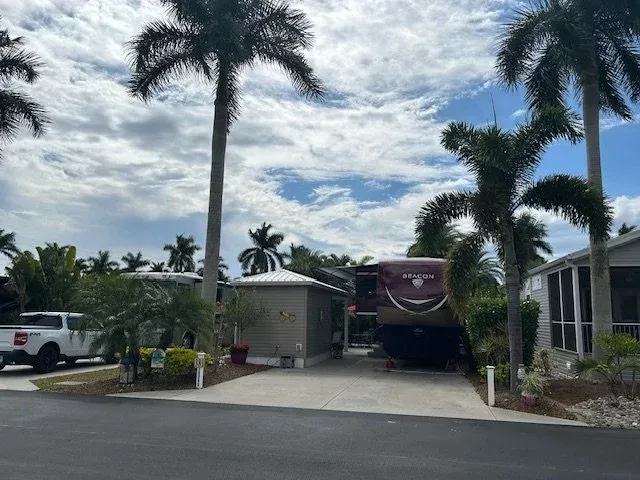 RV parked in a driveway next to a small house, palm trees overhead, under a cloudy sky.