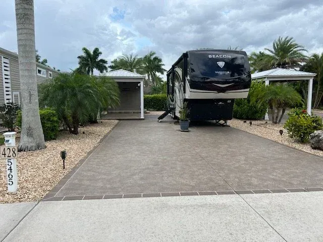 RV parked in a driveway with two small garages. Palm trees, brown driveway.