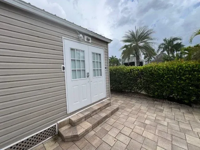 Exterior view of a small beige building with white double doors, brick patio, and green hedge under a cloudy sky.