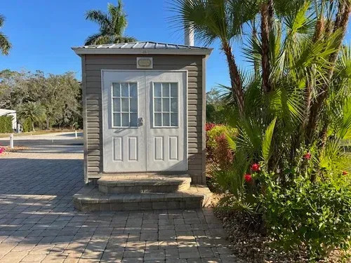 Small gray building with double doors, stone steps, and surrounding landscaping.
