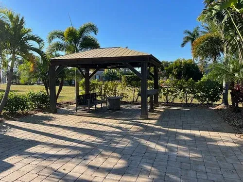 Gazebo on a brick patio, surrounded by green bushes and palm trees under a clear blue sky.