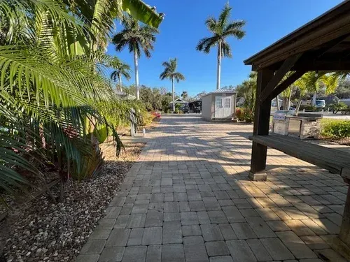 Brick path with gazebo in foreground, palm trees, and small building under blue sky.