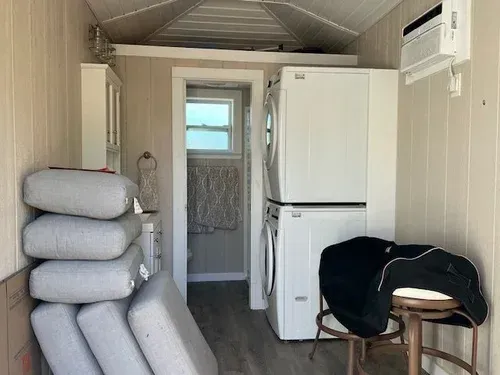 Interior of a small building with stacked washer/dryer, bathroom, cushions, and a saddle on a stool.